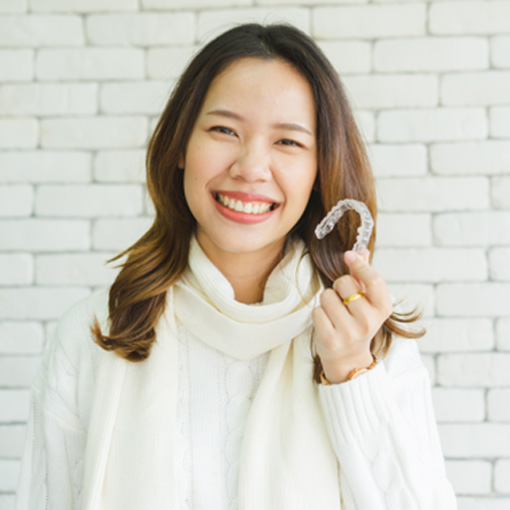 Woman in white sweater in front of white brick wall smiling holding clear aligner